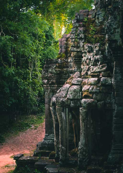 A historical stone temple ruin, possibly part of Ta Prohm or Angkor Wat, with intricate carvings and moss, bordering a reddish dirt path leading into lush green jungle at Khmoch Gate.