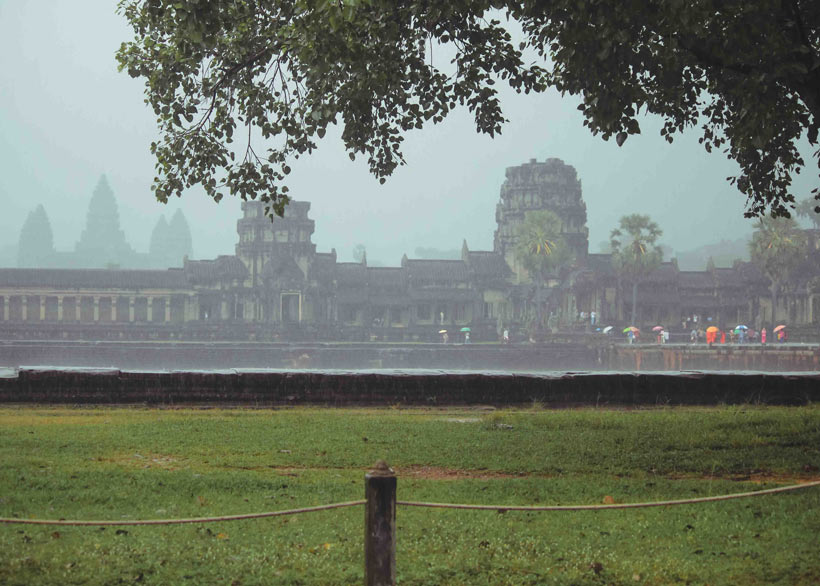Atmospheric view of Angkor Wat during a downpour, highlighting the weathered stone and tropical vegetation.