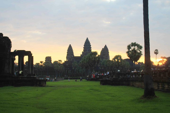 Angkor Wat at sunrise with a glowing sky, green grass, palm trees, and early morning visitors in Siem Reap, Cambodia.