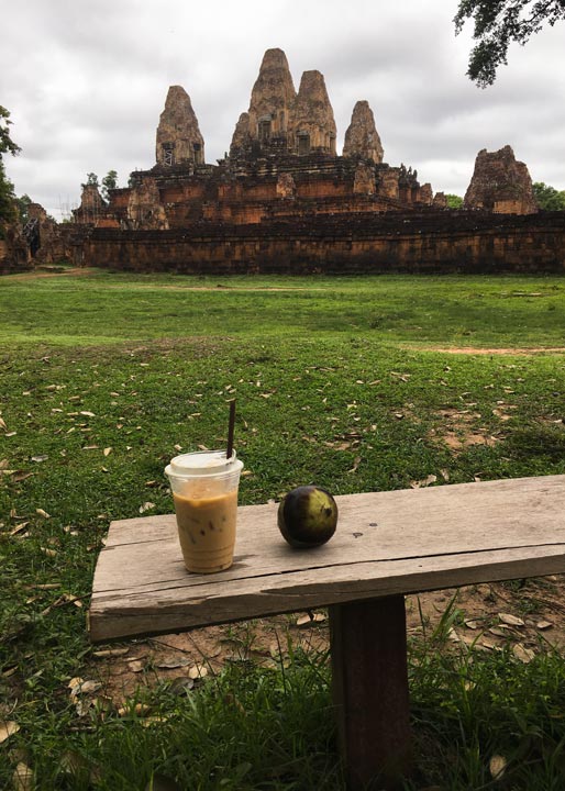 Iced latte and a Palm tree tropical fruit on a wooden bench with a view of Pre Rup Temple under a cloudy sky.