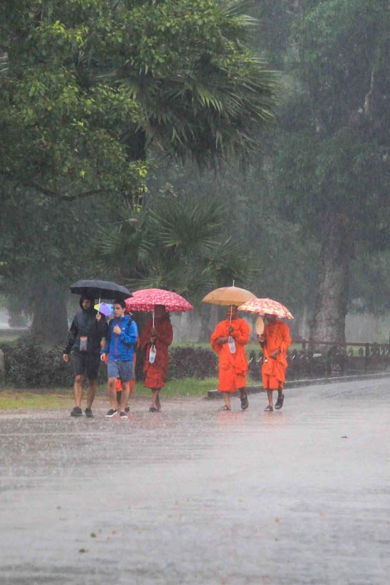 A group of people with umbrellas, some appearing to be Buddhist monks in their traditional orange attire, walk in the rain at Angkor Wat.