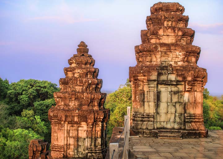 Two ancient temple towers of Phnom Bakheng are bathed in the warm light of the setting sun, with lush green foliage in the background and a clear sky transitioning from blue to purple.