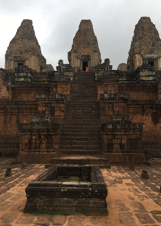 View of the central staircase on the east side of Pre Rup Temple leading up to the main towers, with a cloudy sky and a visitor in red at the upper doorway.