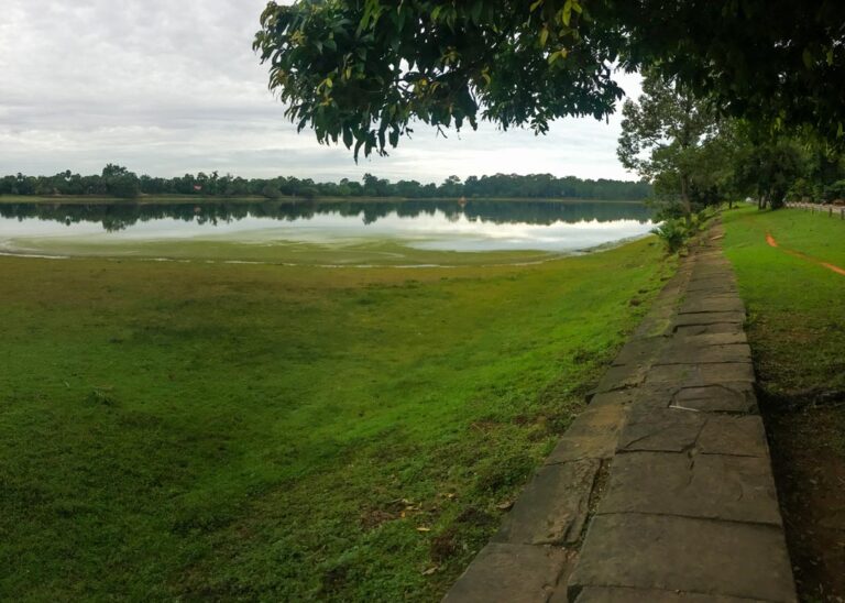 A wide-angle view of the Srah Srang baray, an ancient reservoir, with a stone walkway on the right and lush green grass sloping down to the water's edge. The sky is overcast, and trees line the far bank, reflecting in the calm water.